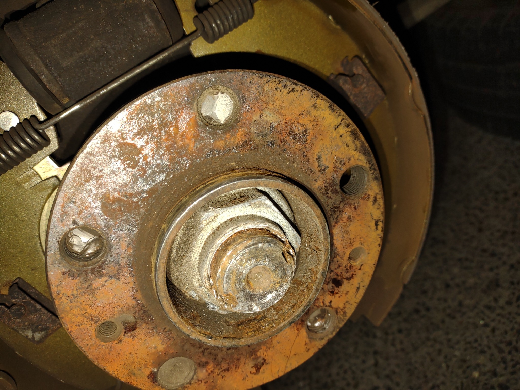 Zardzewiały piasta koła samochodowego z widocznymi śrubami, sprężyną i elementami układu hamulcowego bębnowego.