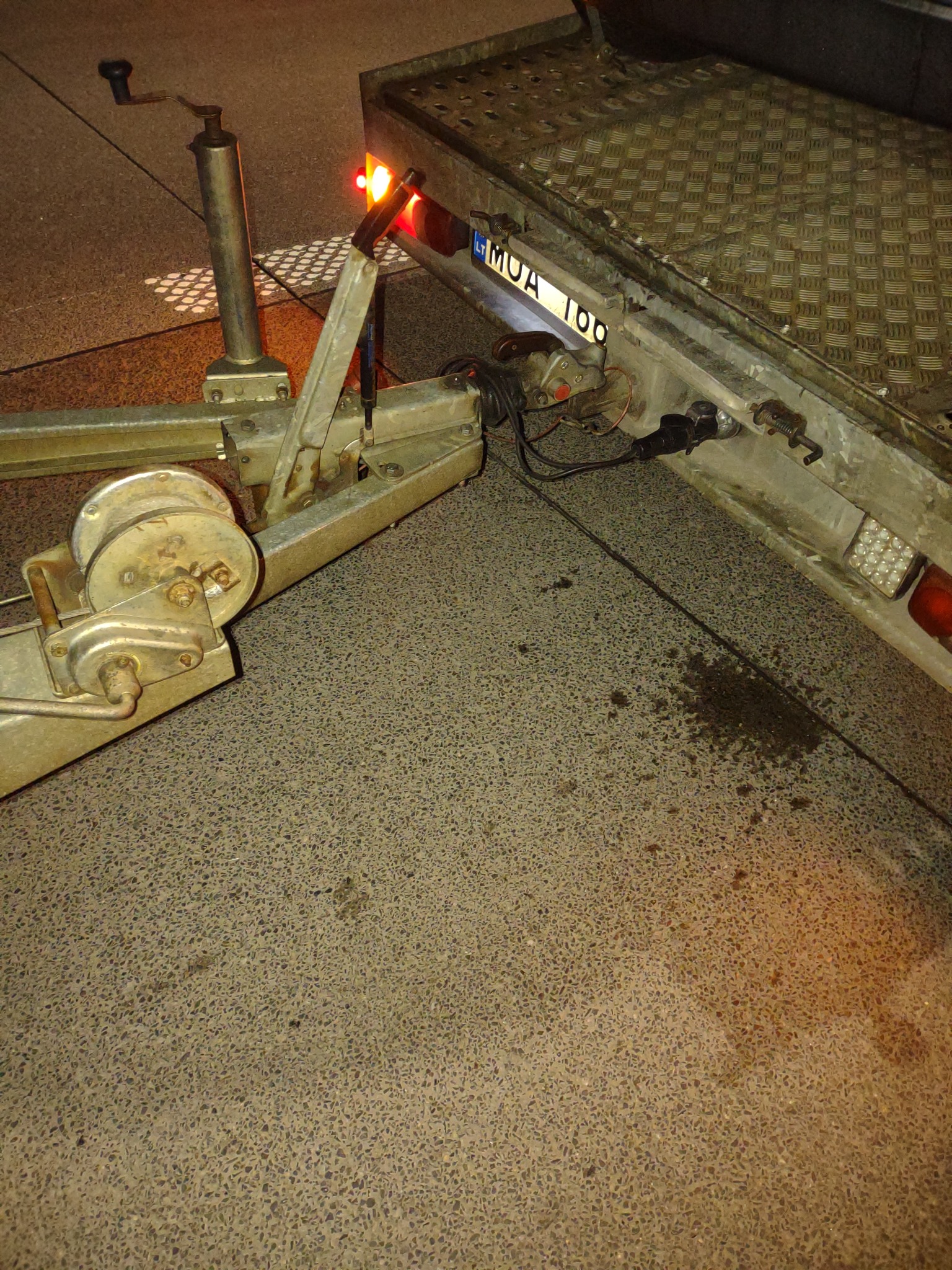 Widok z bliska na metalową lawetę samochodową z widoczną wciągarką, światłem pozycyjnym i tablicą rejestracyjną MUA 100, stojącą na betonowej nawierzchni z plamą oleju.