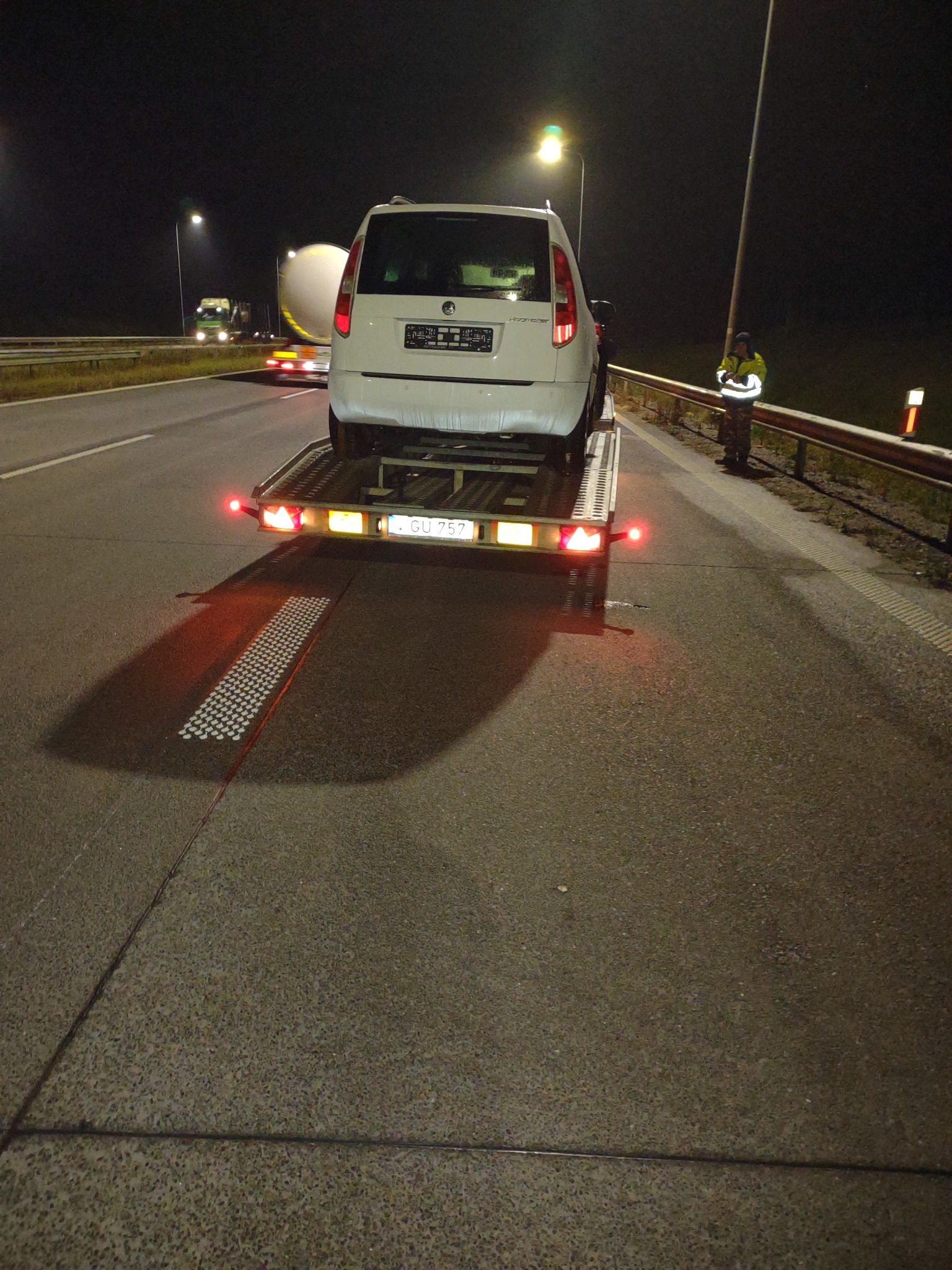 Biały samochód osobowy marki Skoda, transportowany na przyczepie lawetowej na autostradzie nocą, widok z tyłu, oświetlenie drogowe.