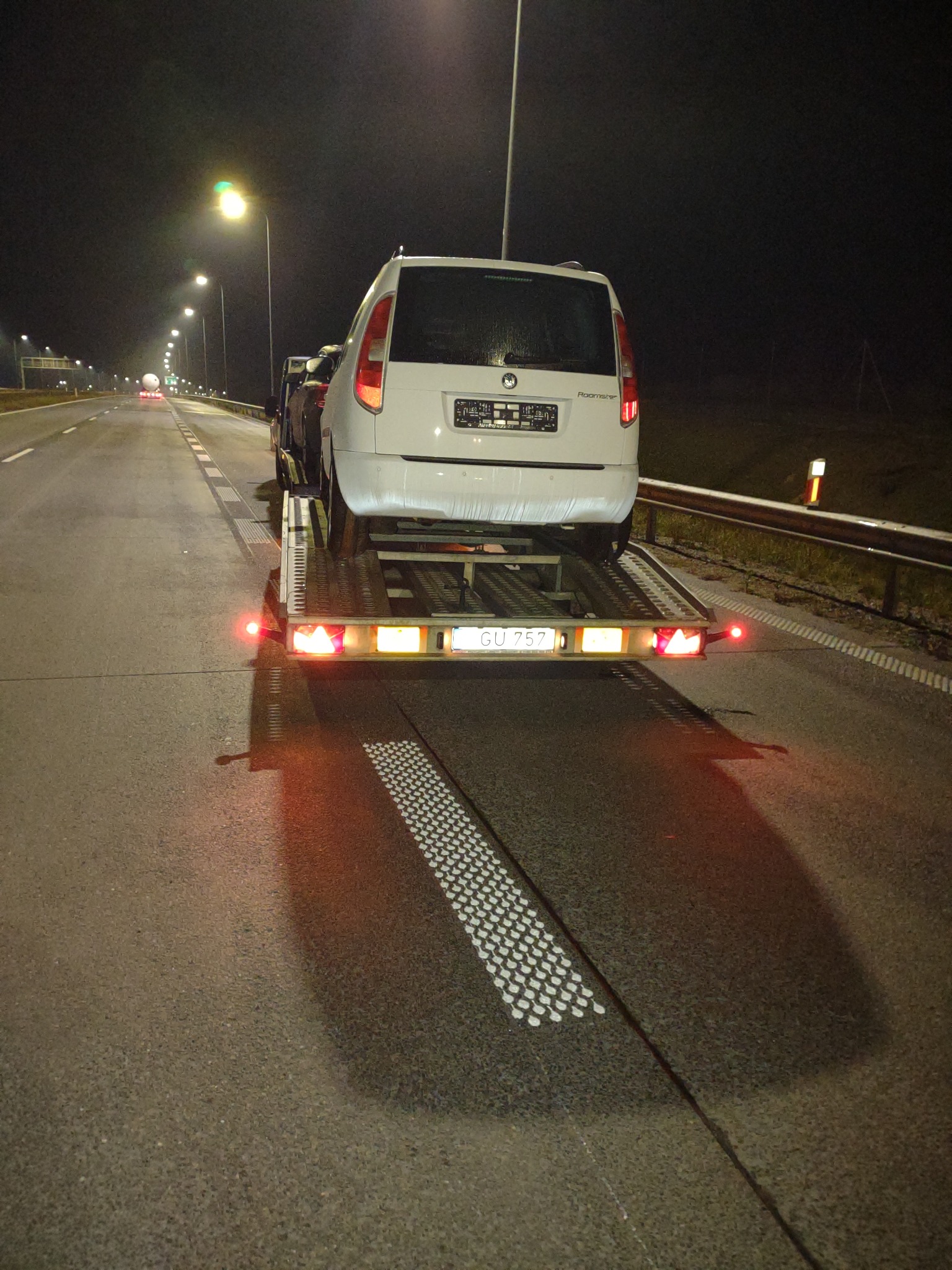 Biały samochód osobowy Skoda Roomster transportowany na lawecie w nocy, widok z tyłu na oświetlonej drodze ekspresowej.