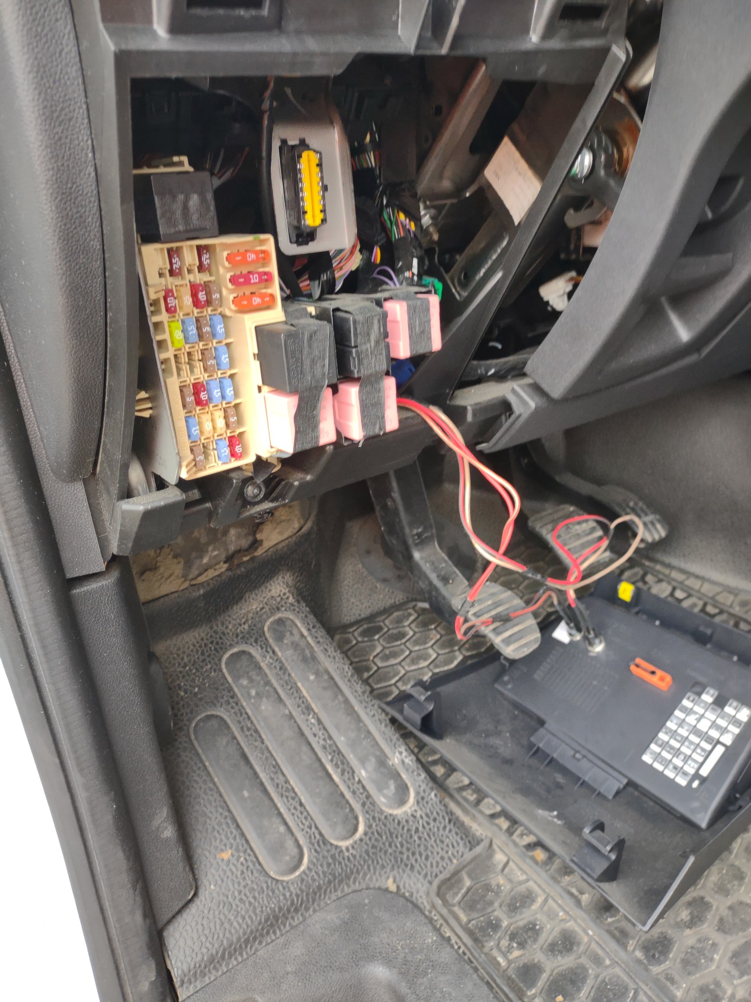 Otwarta skrzynka bezpieczników z widocznymi bezpiecznikami i przekaźnikami w samochodzie w Bydgoszczy, zdemontowana osłona leży na podłodze. Widoczne przewody i pedały.