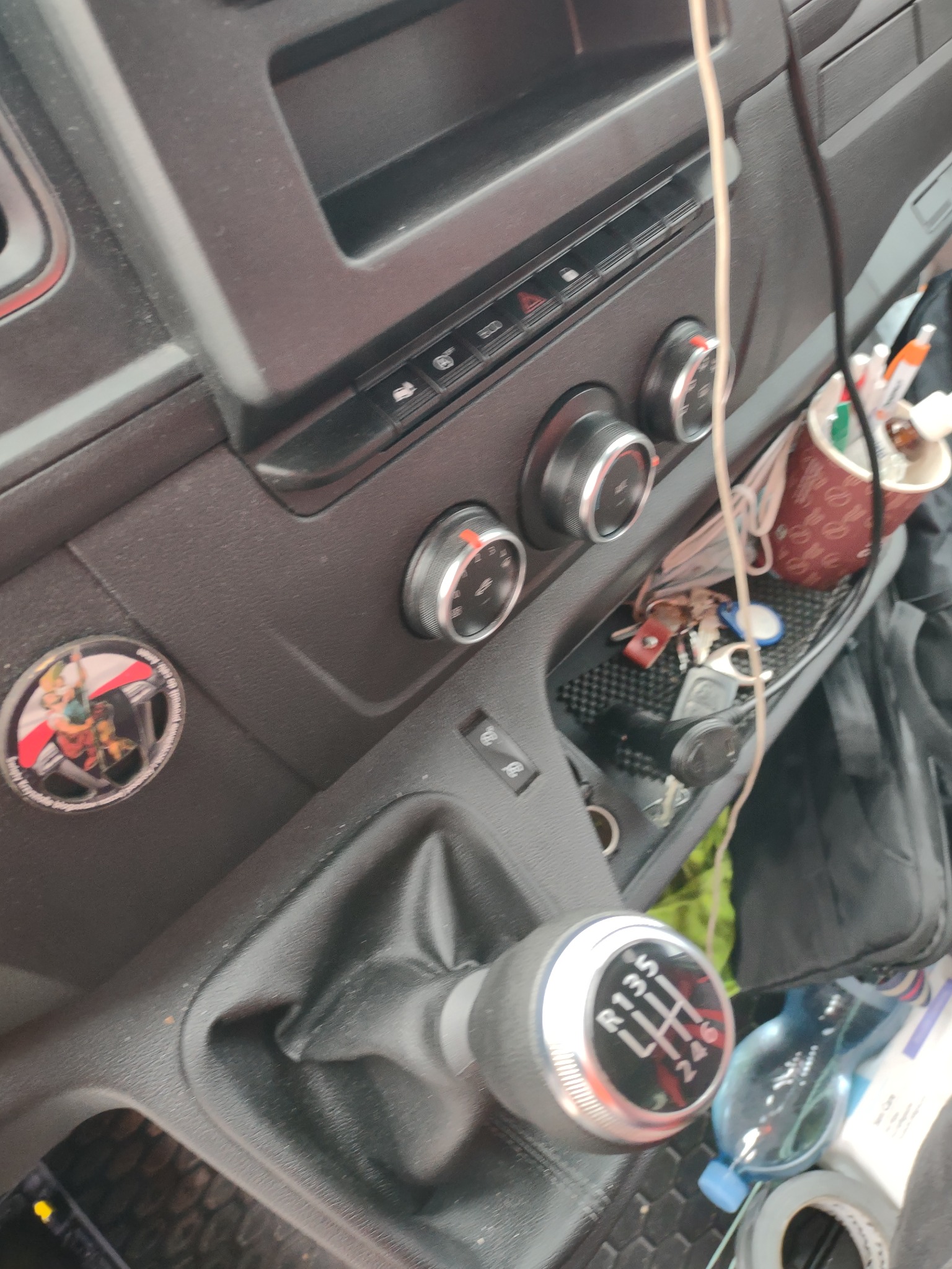 Wnętrze kabiny busa z manualną skrzynią biegów, widoczna gałka zmiany biegów, panel sterowania, kubek z długopisami, klucze i butelka wody.