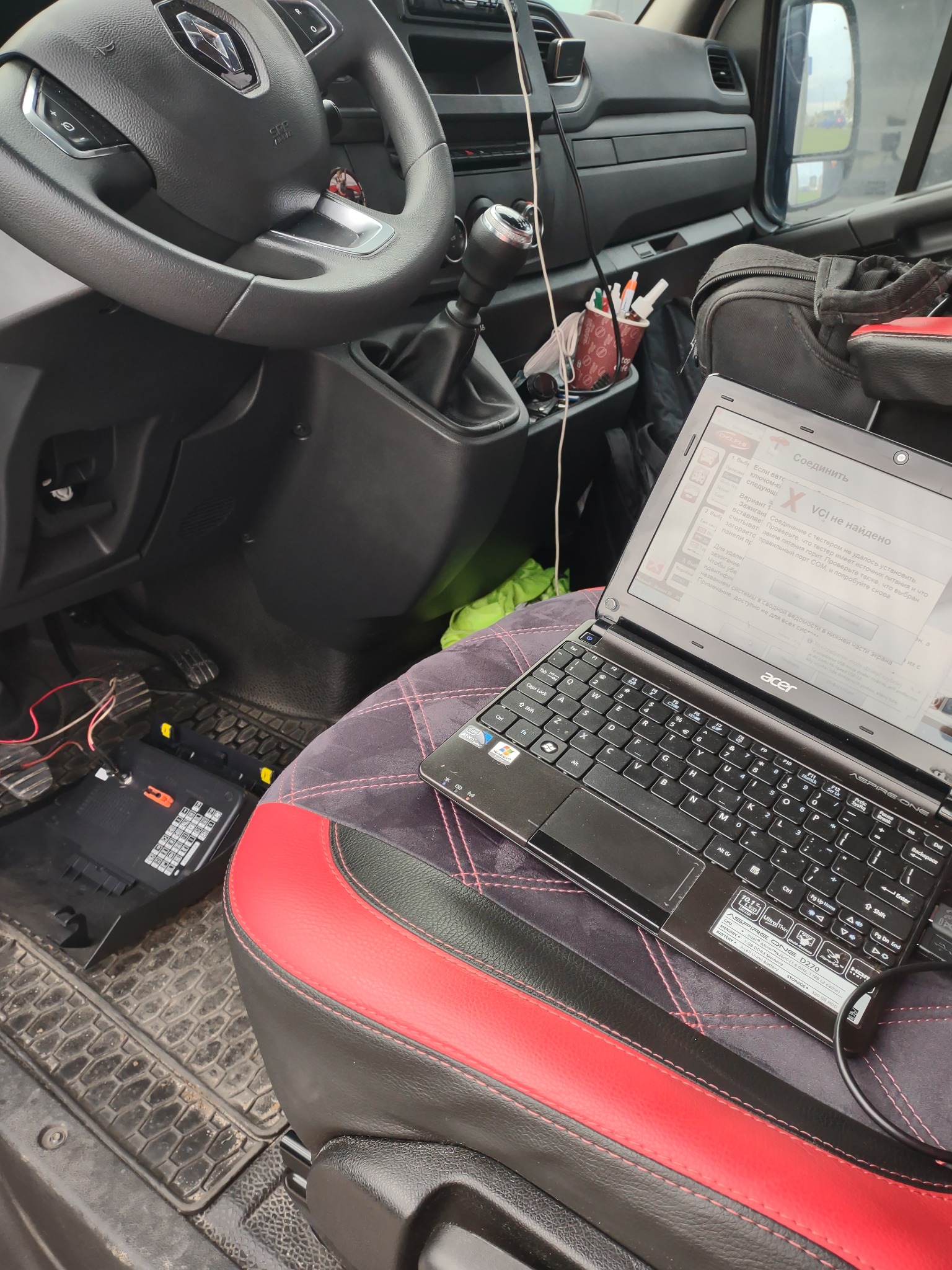 Wnętrze busa: laptop Acer Aspire One na fotelu pasażera z czerwono-czarną tapicerką, podłączony do instalacji elektrycznej pojazdu; diagnostyka samochodowa w toku, widoczny bezpiecznik i kable.
