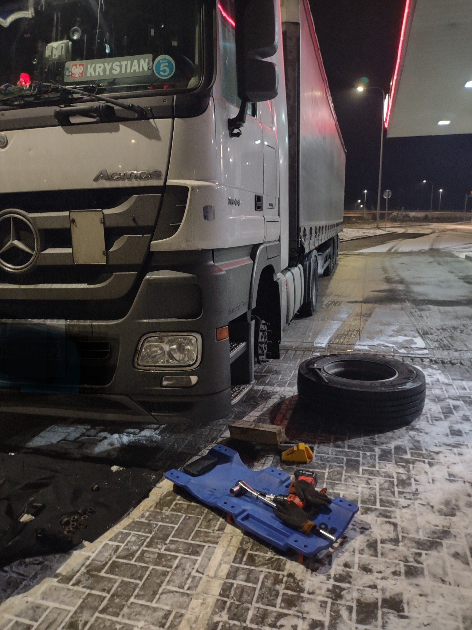 Ciężarówka Mercedes Actros 1844 z odkręconym kołem na stacji benzynowej, obok leży opona, narzędzia i podkładka warsztatowa.