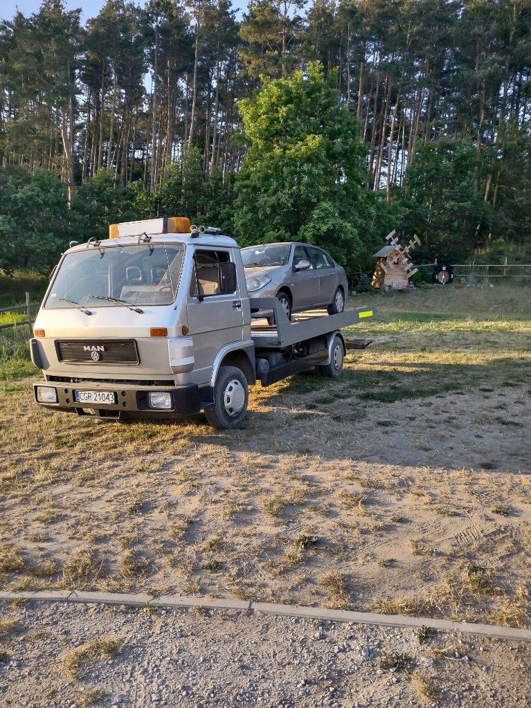 Szara laweta MAN z zaparkowanym szarym samochodem osobowym na platformie, na tle drzew i drewnianego wiatraka.