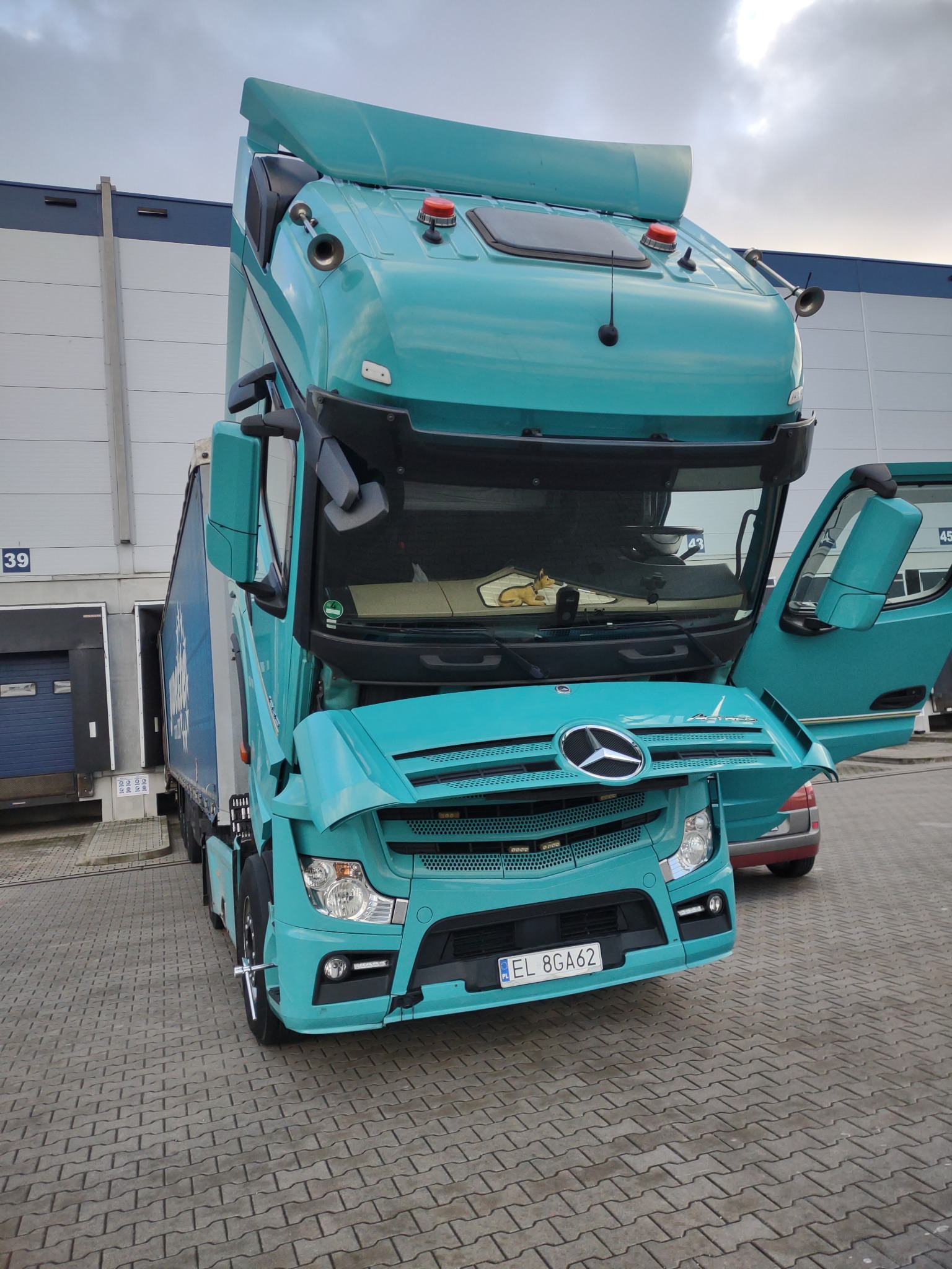 Turkusowy ciągnik siodłowy Mercedes Actros z otwartą kabiną i drzwiami, zaparkowany przed magazynem z numerem 39, widoczny fragment naczepy i tablica rejestracyjna EL 8GA62.