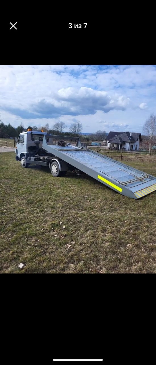 Szara laweta z rozłożoną platformą na trawiastym terenie, w tle dom jednorodzinny i las