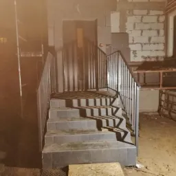 Schody zewnętrzne z metalową balustradą w surowym stanie, widoczne niedokończone ściany budynku i elementy konstrukcyjne.