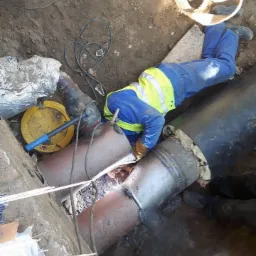 Spawacz w niebieskim kombinezonie i żółtej kamizelce łączy rury w wykopie, widoczne iskry i sprzęt spawalniczy, ziemia wokół.