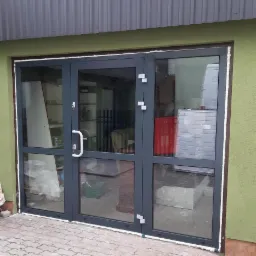 Szare, trzyskrzydłowe drzwi szklane z aluminiową ramą, widoczne z zewnątrz budynku z zieloną elewacją i szarą blachą falistą.