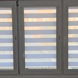 Oknosfera Tomasz Łapiński - Trzyczęściowe okno z białymi ramami i zamontowanymi żaluzjami dzień-noc w poziome pasy, widok z wnętrza pomieszczenia na budynki za oknem.