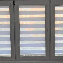 Trzyczęściowe okno z białymi ramami i zamontowanymi żaluzjami dzień-noc w poziome pasy, widok z wnętrza pomieszczenia na budynki za oknem.