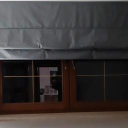 Oknosfera Tomasz Łapiński - Szary materiałowy rolet rzymski na oknie z drewnianą ramą i złotymi szprosami.