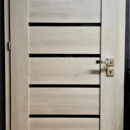 Nowoczesne drzwi wewnętrzne z jasnego drewna z poziomymi, czarnymi wstawkami i minimalistyczną, srebrną klamką.