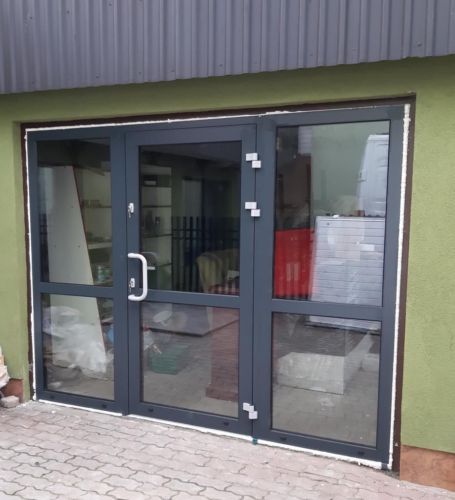 Szare, trzyskrzydłowe drzwi szklane z aluminiową ramą, widoczne z zewnątrz budynku z zieloną elewacją i szarą blachą falistą.