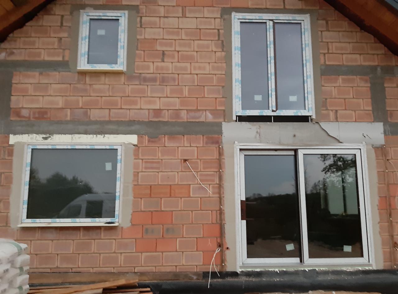 Murowany budynek w trakcie montażu okien z białymi ramami, widoczne ślady zaprawy i wystające przewody.