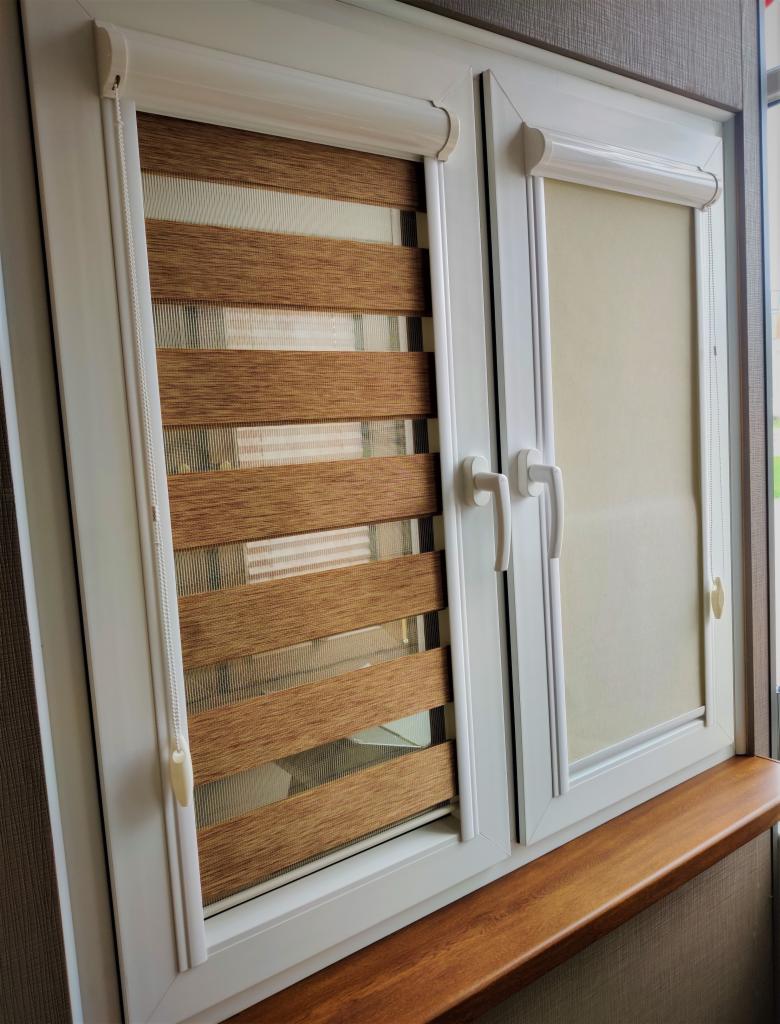 Białe okno PCV z brązowymi żaluzjami dzień-noc po lewej stronie i beżową roletą po prawej, zamontowane na drewnianym parapecie.