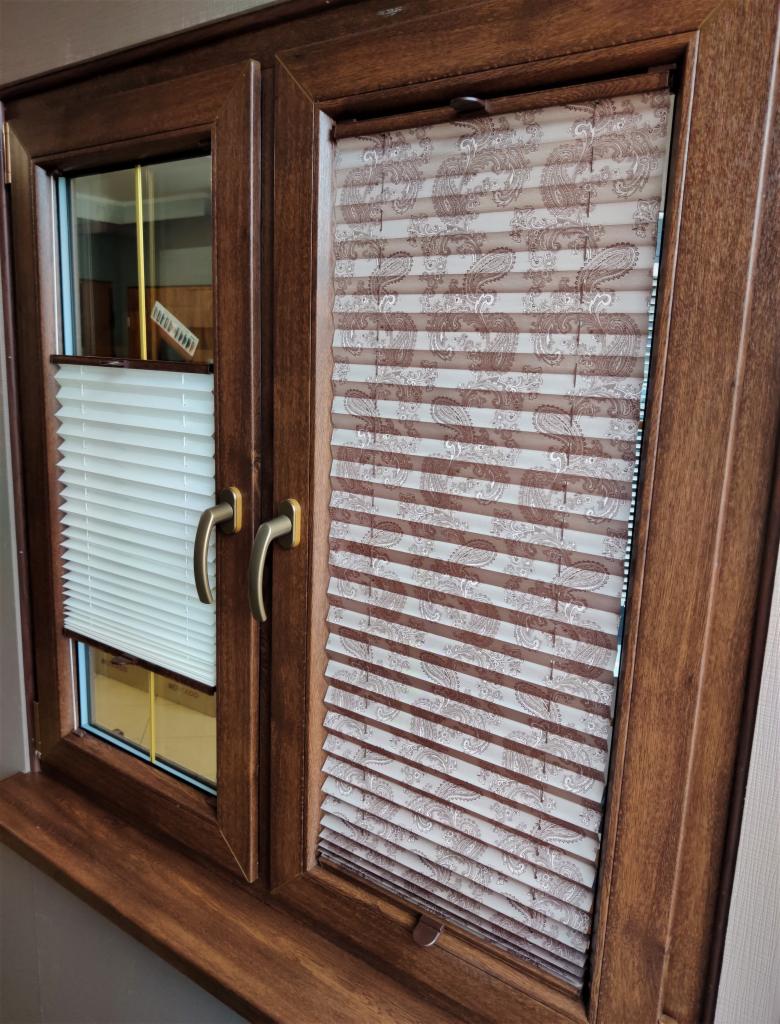Drewniane okno PCV z dwiema częściami: jedna z plisowaną żaluzją w kolorze białym, a druga z żaluzją plisowaną z brązowym wzorem paisley.