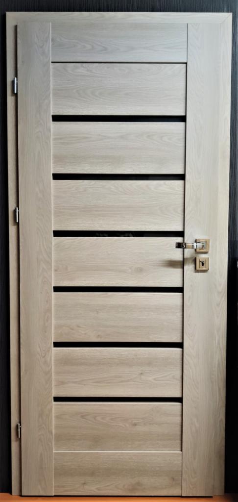Nowoczesne drzwi wewnętrzne z jasnego drewna z poziomymi, czarnymi wstawkami i minimalistyczną, srebrną klamką.