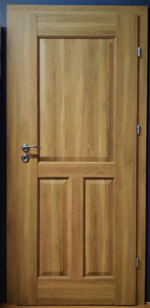 Drewniane drzwi wewnętrzne z trzema panelami i srebrną klamką na tle ciemnej ściany.