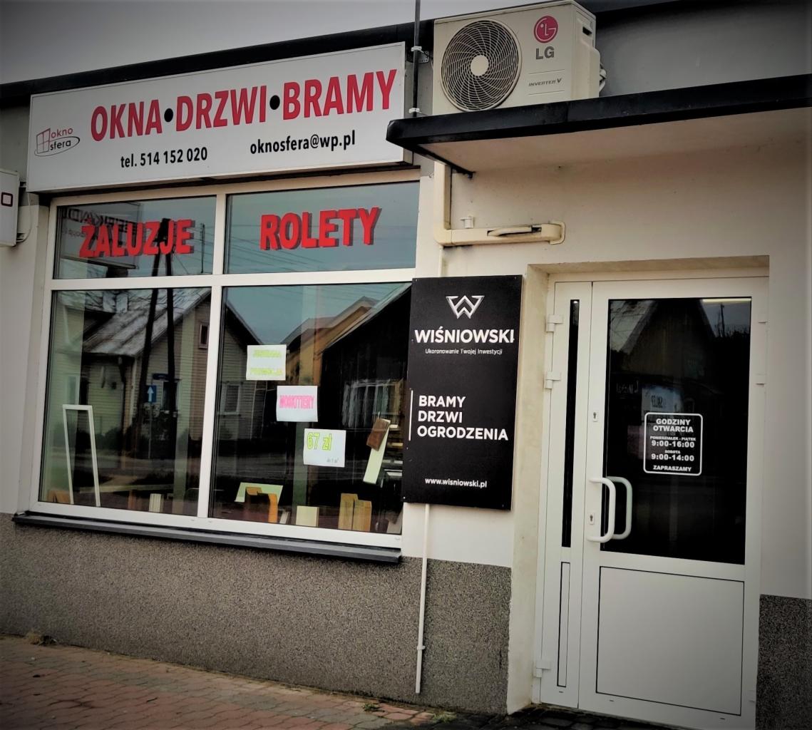 Fasada budynku firmy oferującej okna, drzwi i bramy z widocznym szyldem, oknami wystawowymi z ekspozycją żaluzji i rolet oraz tablicą informacyjną marki Wiśniowski.
