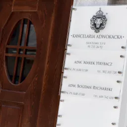 Tablica informacyjna Kancelarii Adwokackiej z logo lwa w koronie, adres Mostowa 13/4 i nazwiskami adwokatów Marka Hrybacza i Bogdana Rycharskiego obok drewnianych drzwi z owalnym okienkiem.