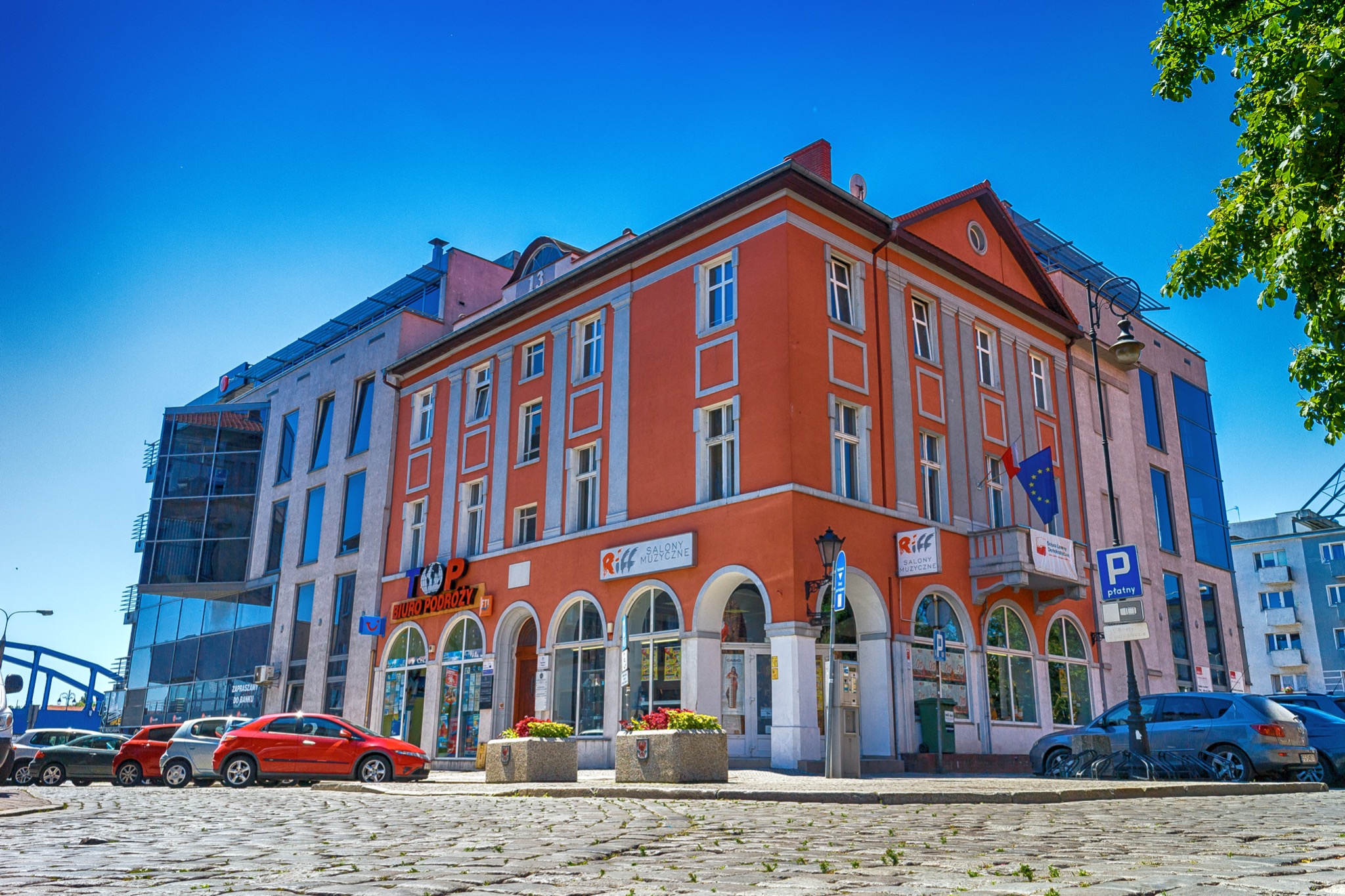 Elewacja kamienicy z biurami i salonami usługowymi, widok z poziomu brukowanej ulicy w słoneczny dzień.