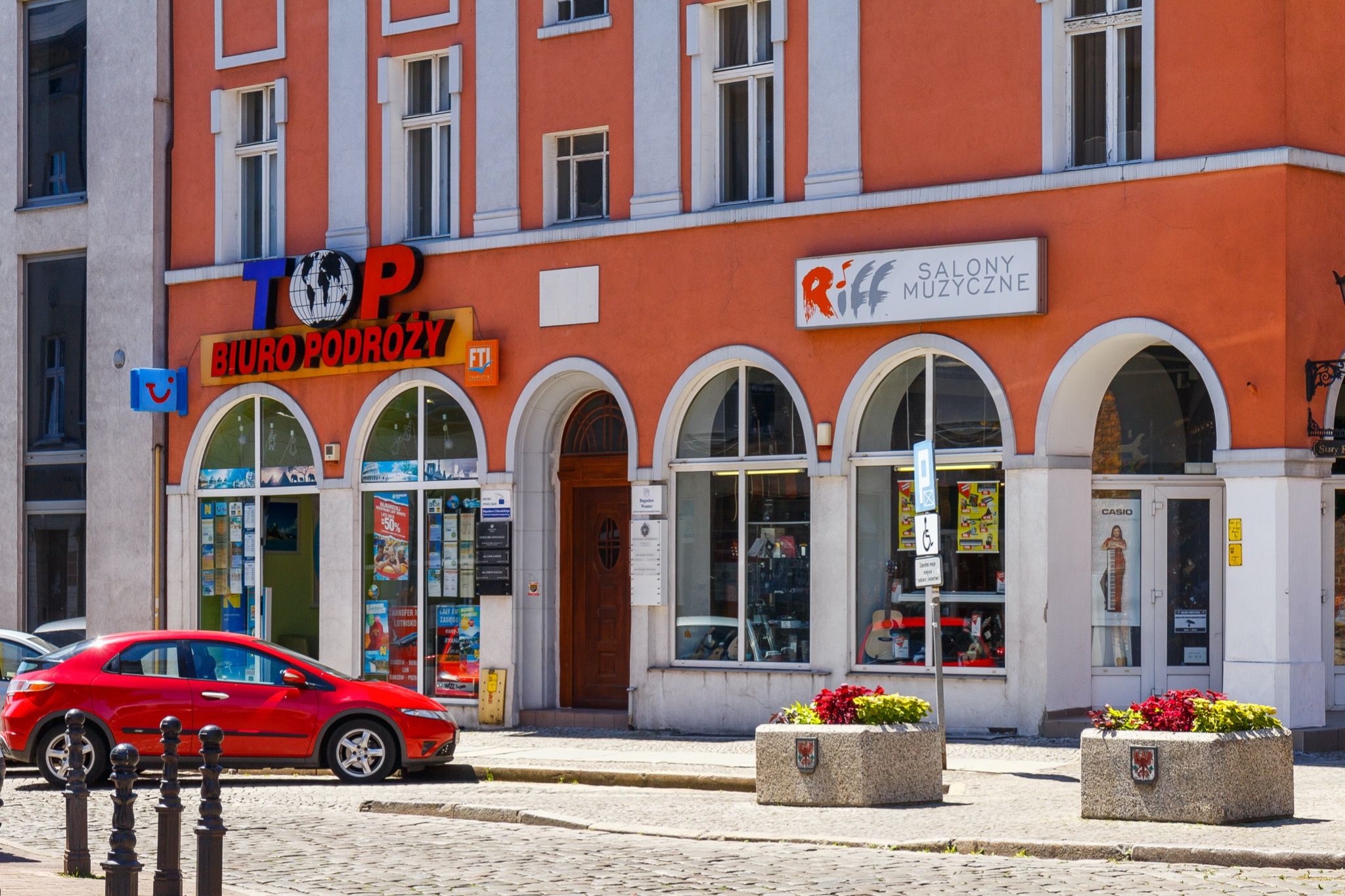 Fasada pomarańczowego budynku z białymi obramowaniami okien i łukami nad wejściami, mieszczącego Biuro Podróży TOP z globusem w logo oraz Salon Muzyczny Riff, z czerwonym samochodem zaparkowanym...