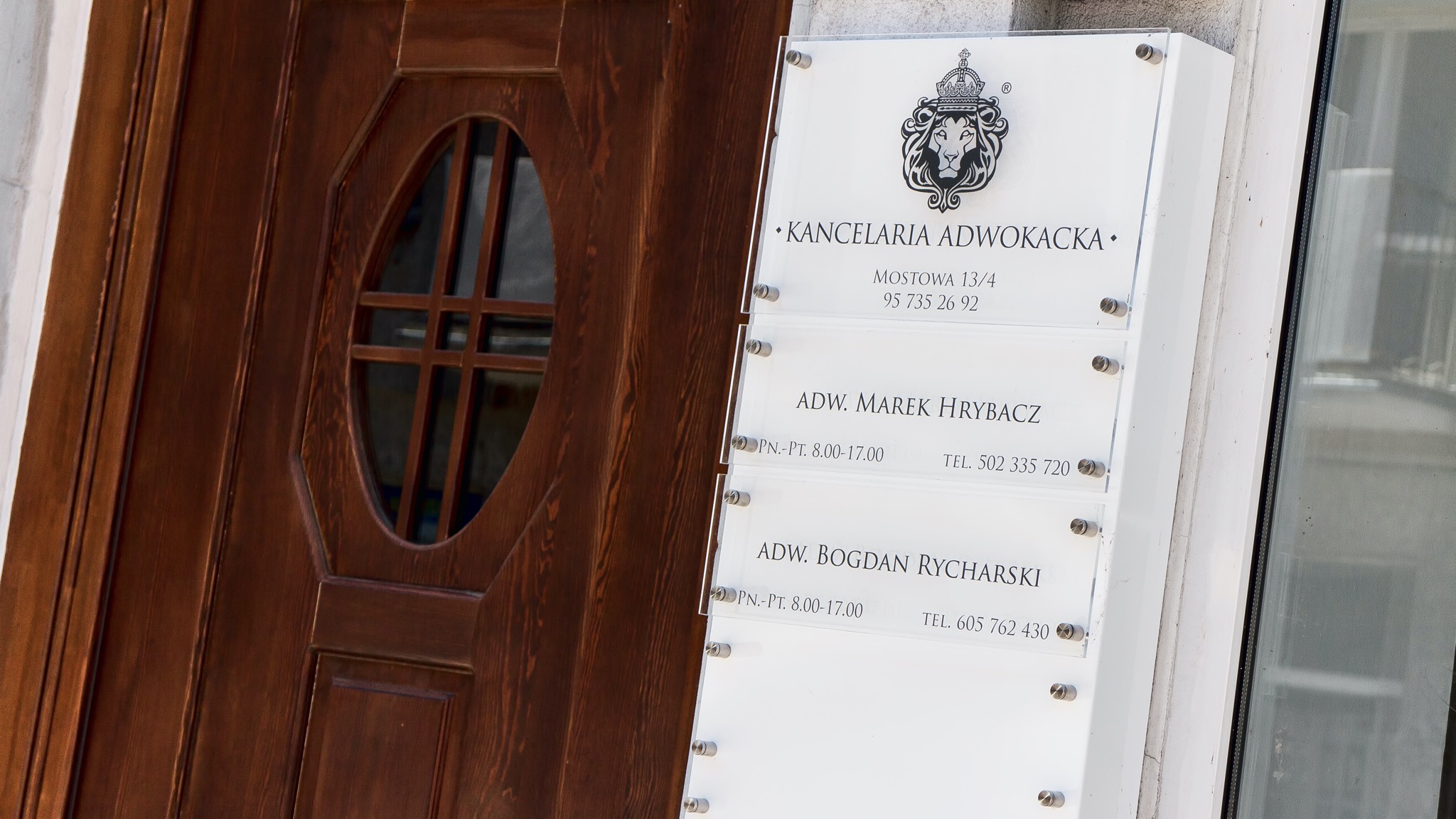 Tablica informacyjna Kancelarii Adwokackiej z logo lwa w koronie, adres Mostowa 13/4 i nazwiskami adwokatów Marka Hrybacza i Bogdana Rycharskiego obok drewnianych drzwi z owalnym okienkiem.