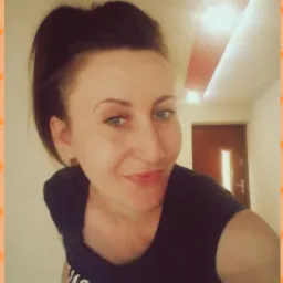 Portret uśmiechniętej kobiety w Brzezinach, z włosami upiętymi w wysoki kucyk, w czarnej koszulce z napisem, na tle jasnego korytarza z brązowymi drzwiami.