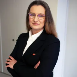 Portret kobiety w okularach, czarnej marynarce i białej koszuli, z rękami skrzyżowanymi, na tle białych drzwi i paneli ściennych.