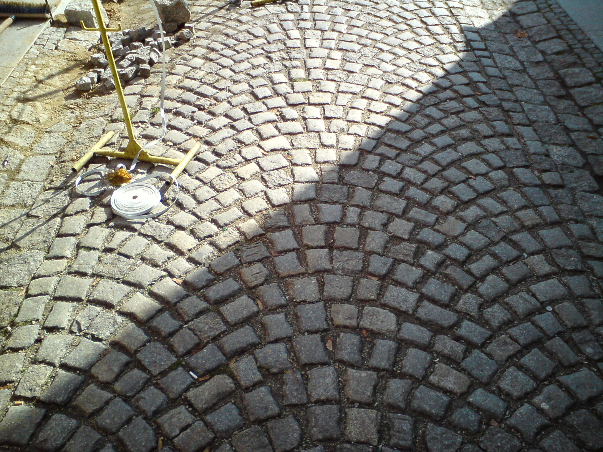 Półokrągły wzór z szarej kostki brukowej, częściowo zacieniony, z żółtym statywem i rolką taśmy w lewym górnym rogu.