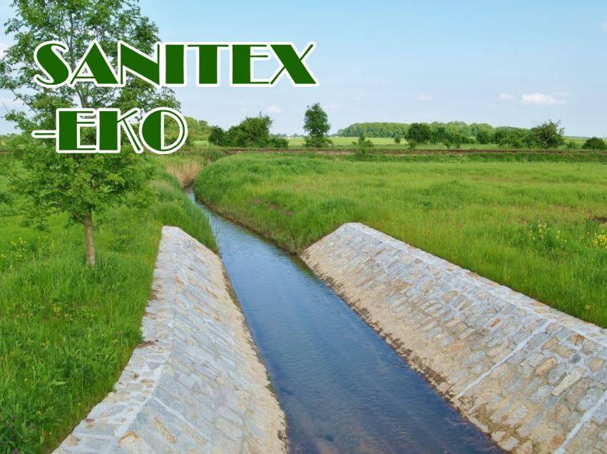 Ujęcie z góry na kanał z wodą, wyłożony kamieniami, otoczony zielenią. W tle widoczne drzewa i błękitne niebo. Logo firmy SANITEX-EKO w lewym górnym rogu.