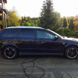 Ciemny samochód kombi Audi A4 B6 po umyciu, stojący na kostce brukowej, z widocznym wężem myjki ciśnieniowej i budynkiem w tle, w promieniach zachodzącego słońca.