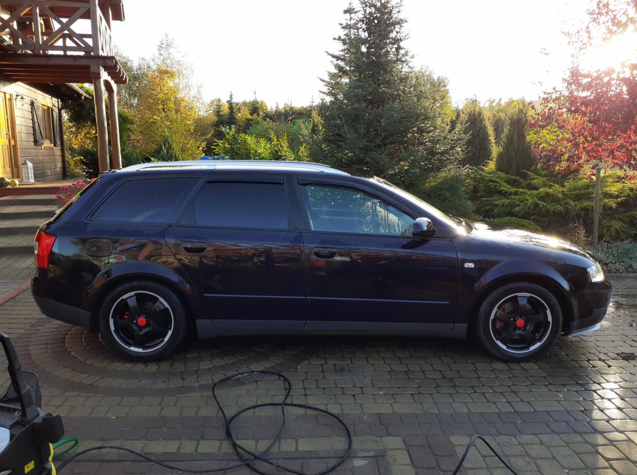 Ciemny samochód kombi Audi A4 B6 po umyciu, stojący na kostce brukowej, z widocznym wężem myjki ciśnieniowej i budynkiem w tle, w promieniach zachodzącego słońca.