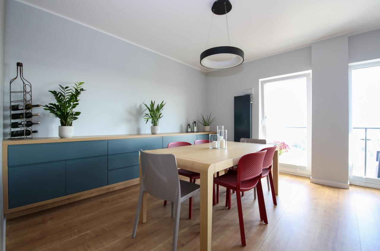 Jasne wnętrze jadalni z drewnianym stołem otoczonym czerwonymi i szarymi krzesłami, nad którym wisi okrągła lampa. W tle komoda z roślinami w doniczkach i metalowy stojak na wino.