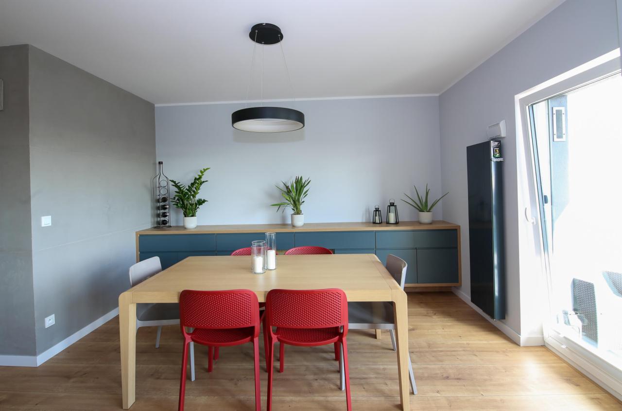 Jasne wnętrze jadalni z drewnianym stołem, czerwonymi krzesłami i minimalistycznym oświetleniem, widok na przeszklone drzwi balkonowe.