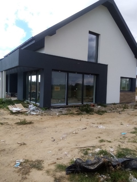 Dwukondygnacyjny dom z szarą elewacją i dużymi przesuwnymi oknami, widoczne ślady prac budowlanych na zewnątrz budynku.