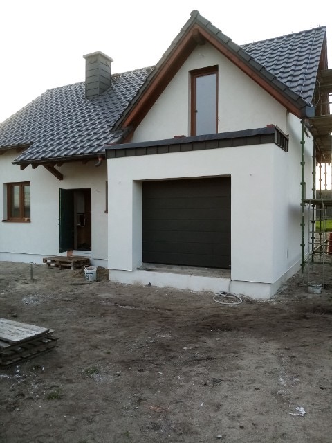 Biały dom jednorodzinny z ciemnoszarą bramą garażową i rusztowaniem z boku, teren wokół domu w trakcie prac wykończeniowych.