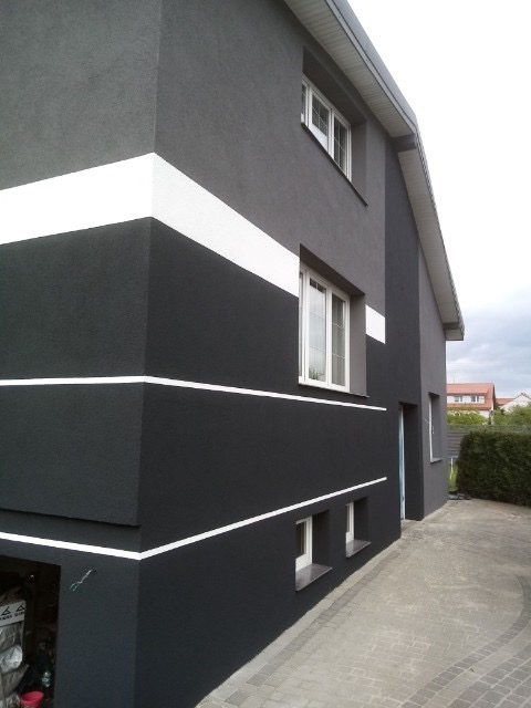 Dwukondygnacyjny dom z ciemnoszarą elewacją ozdobioną białymi poziomymi pasami, widok z boku na garaż i podjazd z kostki brukowej.