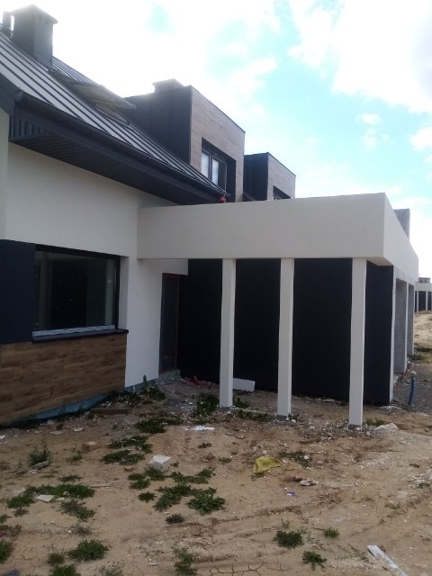 Nowoczesny dom w trakcie budowy z elementami wykończenia elewacji: białe ściany, czarne panele i drewnopodobna okładzina. Widoczne białe kolumny podtrzymujące zadaszenie oraz niezagospodarowany...