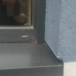 Szczegółowe ujęcie narożnika okna z szarą ramą i parapetem zewnętrznym, przylegającego do niebieskiej, chropowatej ściany elewacyjnej i białej ściany poniżej.