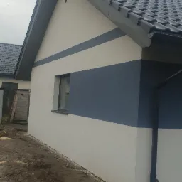 Nowoczesny dom jednorodzinny z biało-granatową elewacją, widok z boku na fragment budynku z oknem i rynną, na pierwszym planie błotniste podłoże.