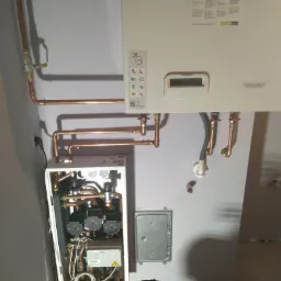 Wnętrze szafki z instalacją grzewczą, widoczne miedziane rury, zawory i podłączenia elektryczne, obok zamontowany na ścianie biały piec grzewczy z wyświetlaczem.