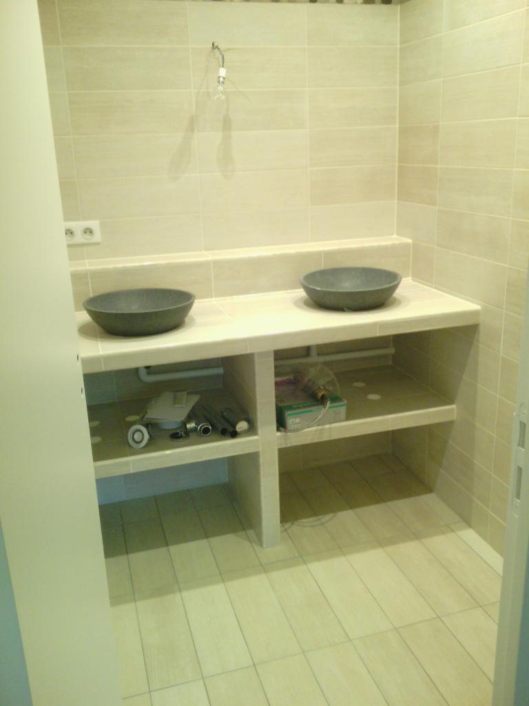 Dwie szare, okrągłe umywalki na jasnym, kafelkowym blacie w łazience, z widocznymi elementami instalacji i narzędziami na półkach poniżej.