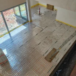 Rozłożone rury ogrzewania podłogowego na folii izolacyjnej z widocznymi punktami mocowania, przygotowane do zalania wylewką, widok z góry na plac budowy.