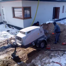 Praca w zimowych warunkach: dwóch mężczyzn obsługuje maszynę do tynkowania/wylewania posadzek na zewnątrz budynku, widoczne worki z cementem i pozostałości śniegu.