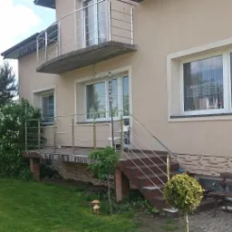 Dom z beżową elewacją, balkonem i wejściem z balustradą ze stali nierdzewnej, widok z ogrodu.