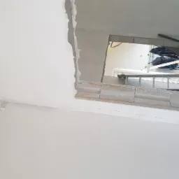 Ściana i sufit po wstępnym wyrównaniu, widoczne ślady szpachli, fragment ściany z pustaków z otworem, drabina w tle.