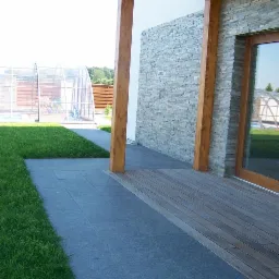 Nowoczesny taras z desek kompozytowych przechodzący w ścieżkę z ciemnej kostki brukowej, z drewnianą pergolą i kamienną ścianą domu w tle, widoczny basen z zadaszeniem.